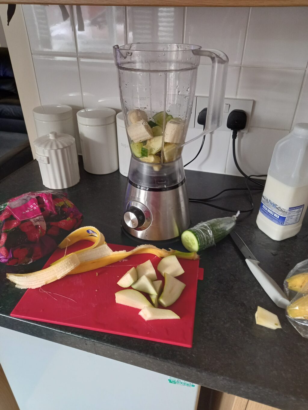 A blender and chopping board containing ingredients to make a smoothie. The blender contains banana and cucumber, and on the chopping board there is more banana and sliced pear. There is also a bag of frozen berries and a bottle of milk on the counter.