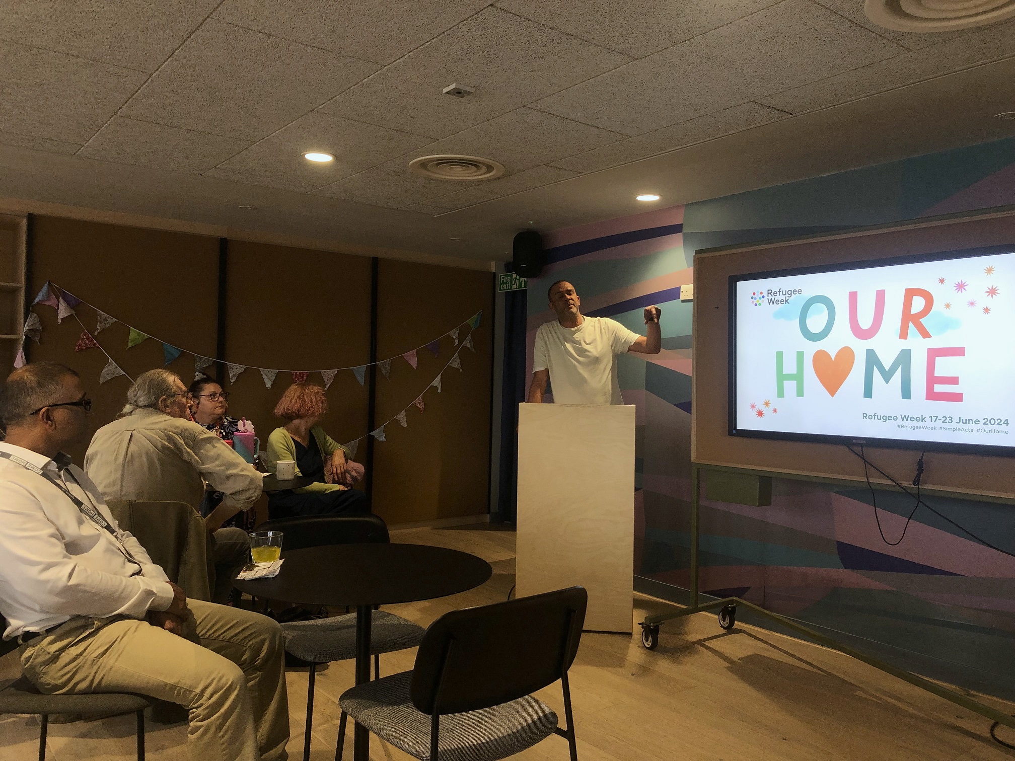 Lewis stands at a lectern next to a screen that says 'Our home'. The room is decorated with bunting and there are people sat around tables in the audience.