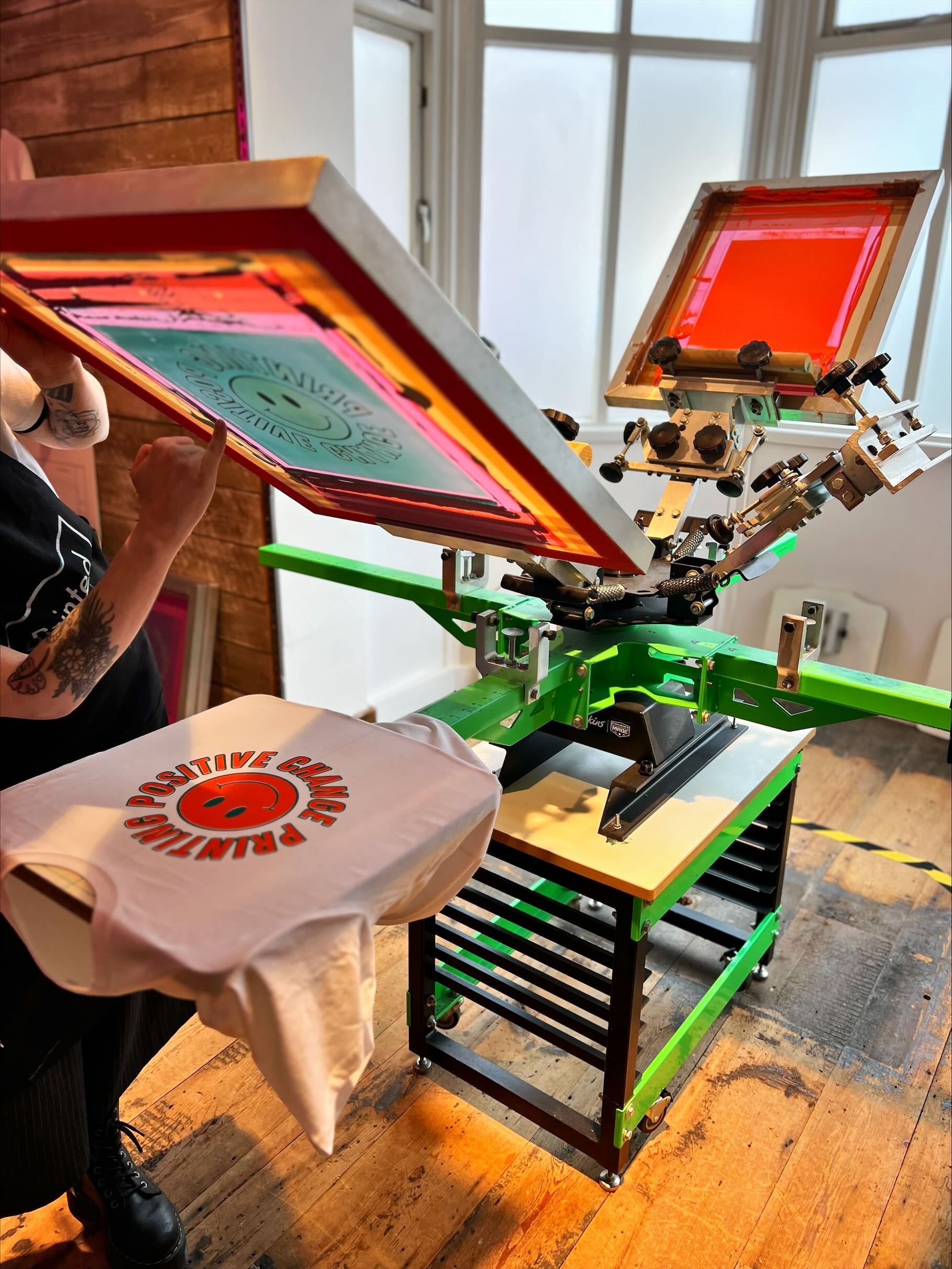 Someone screen-printing a smiley face and the text 'Printing positive change' onto a t-shirt. You can see the large screen-printing machine, the t-shirt, and the person's hands.