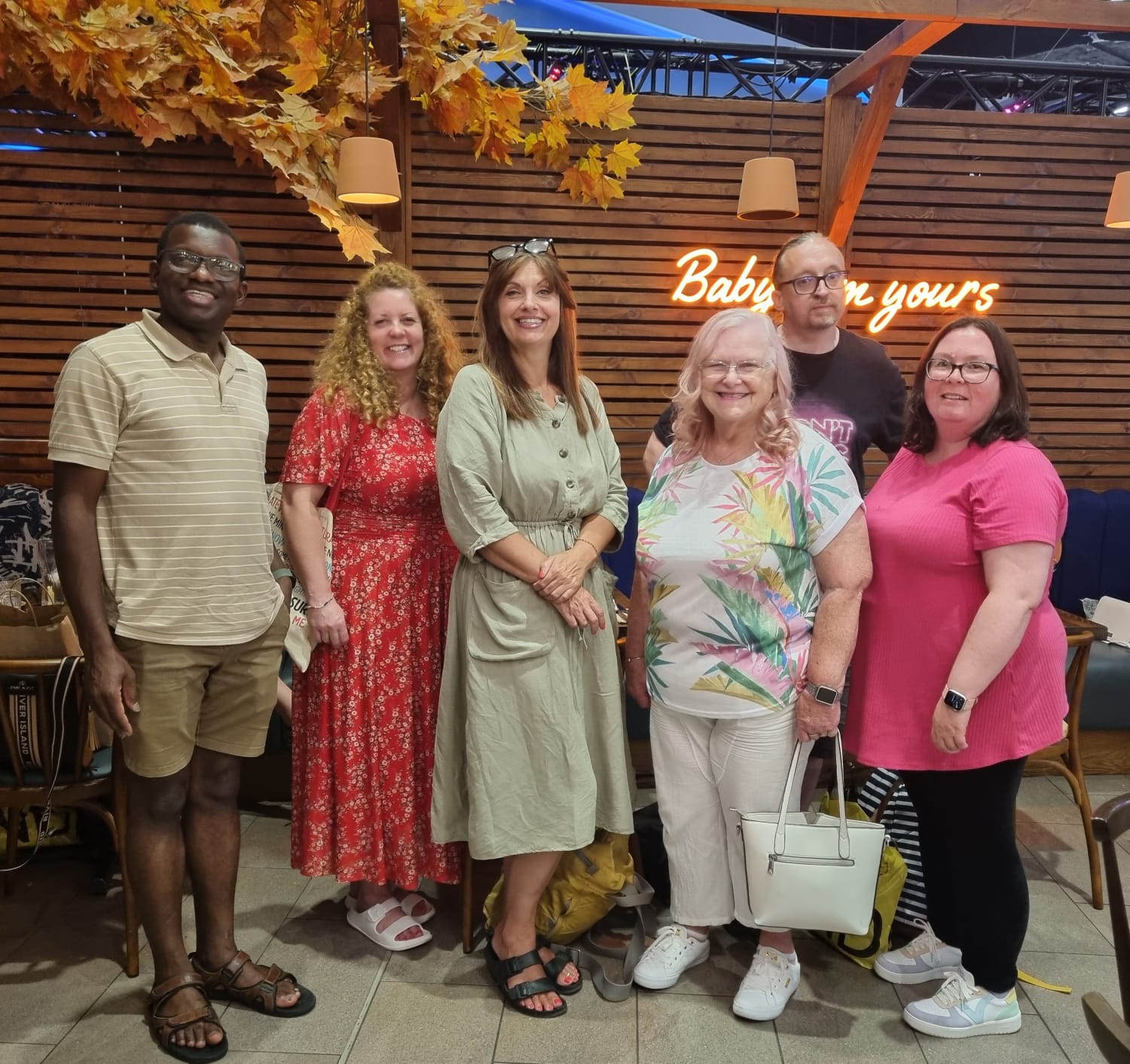 A photo of some of our Customer Experience Team and Customer Scrutiny Panel. Everyone looks very colourful and summery in shorts, sandals and/or bright colours. They are stood in a restaurant and smiling at the camera.